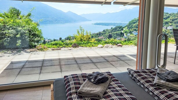 A room with a large glass door leading to a terrace with a scenic view of mountains and a lake. - Nice apartment for 4 guests with WIFI, TV, patio, pets allowed and panoramic view (Tenero)
