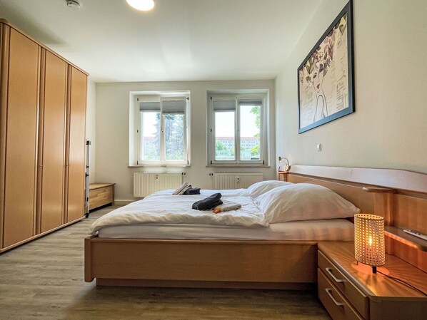 A bedroom with a bed, two windows, a radiator, a wardrobe, a nightstand, and a lamp.