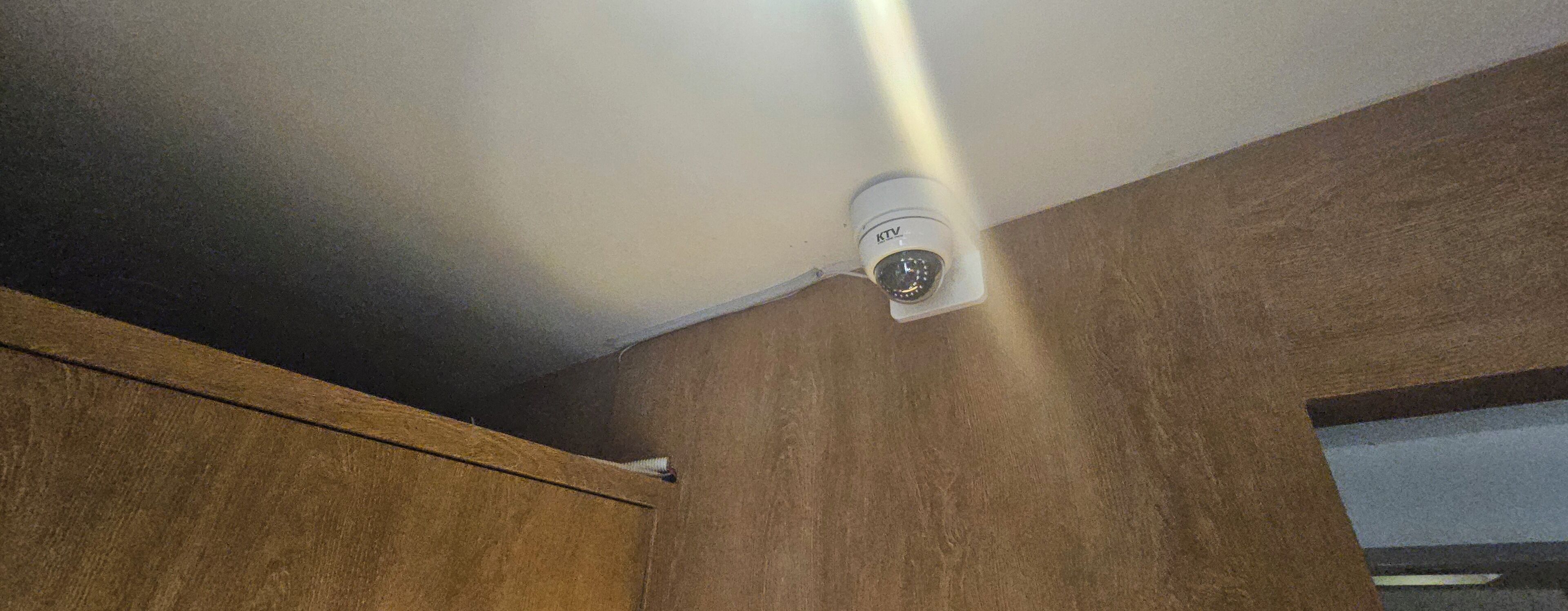 Eine an der Decke befestigte Überwachungskamera in einem Raum mit Holzwänden und einem sichtbaren Türrahmen.