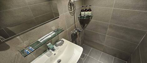 Un baño con repisa de vidrio que contiene artículos de aseo, ducha con cabezal de ducha manual y lavabo blanco con espejo.