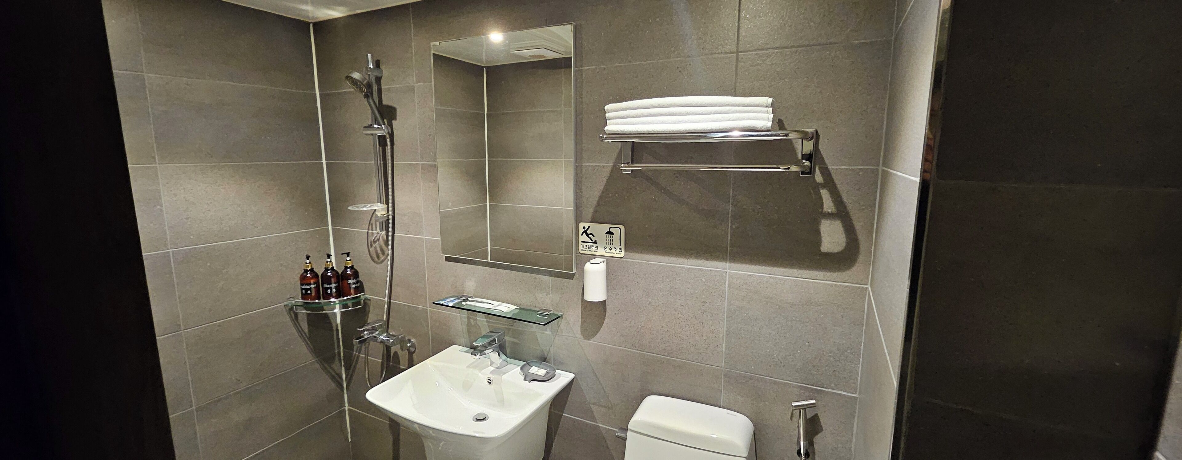 A modern bathroom with a glass shower enclosure, a white sink, and a toilet. There are toiletries on a shelf and a towel rack above the toilet.