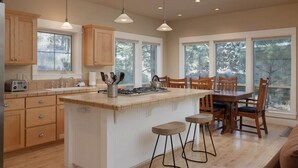 A modern kitchen with wooden cabinets, a central island with stools, and large windows offering views of trees. - Bluebird Day - Big River, Luxurious 4BR home in Bend, perfect for families or groups of up to 8. (Bend)