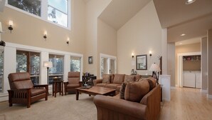 A spacious living room with high ceilings, large windows, and a view of trees. There is a kitchen area visible in the background. - Bluebird Day - Big River, Luxurious 4BR home in Bend, perfect for families or groups of up to 8. (Bend)