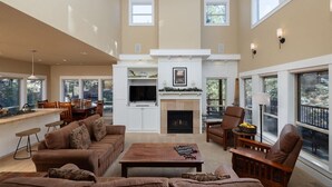 A spacious living room with a fireplace, large windows, and a kitchen area. - Bluebird Day - Big River, Luxurious 4BR home in Bend, perfect for families or groups of up to 8. (Bend)