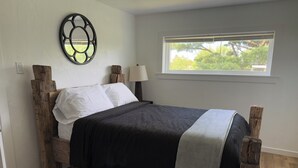 A bedroom with a wooden bed frame, white pillows, a dark bedspread, a window with a view of trees, and a wall-mounted decorative piece. - Farmhouse Fantasy (Nauvoo)