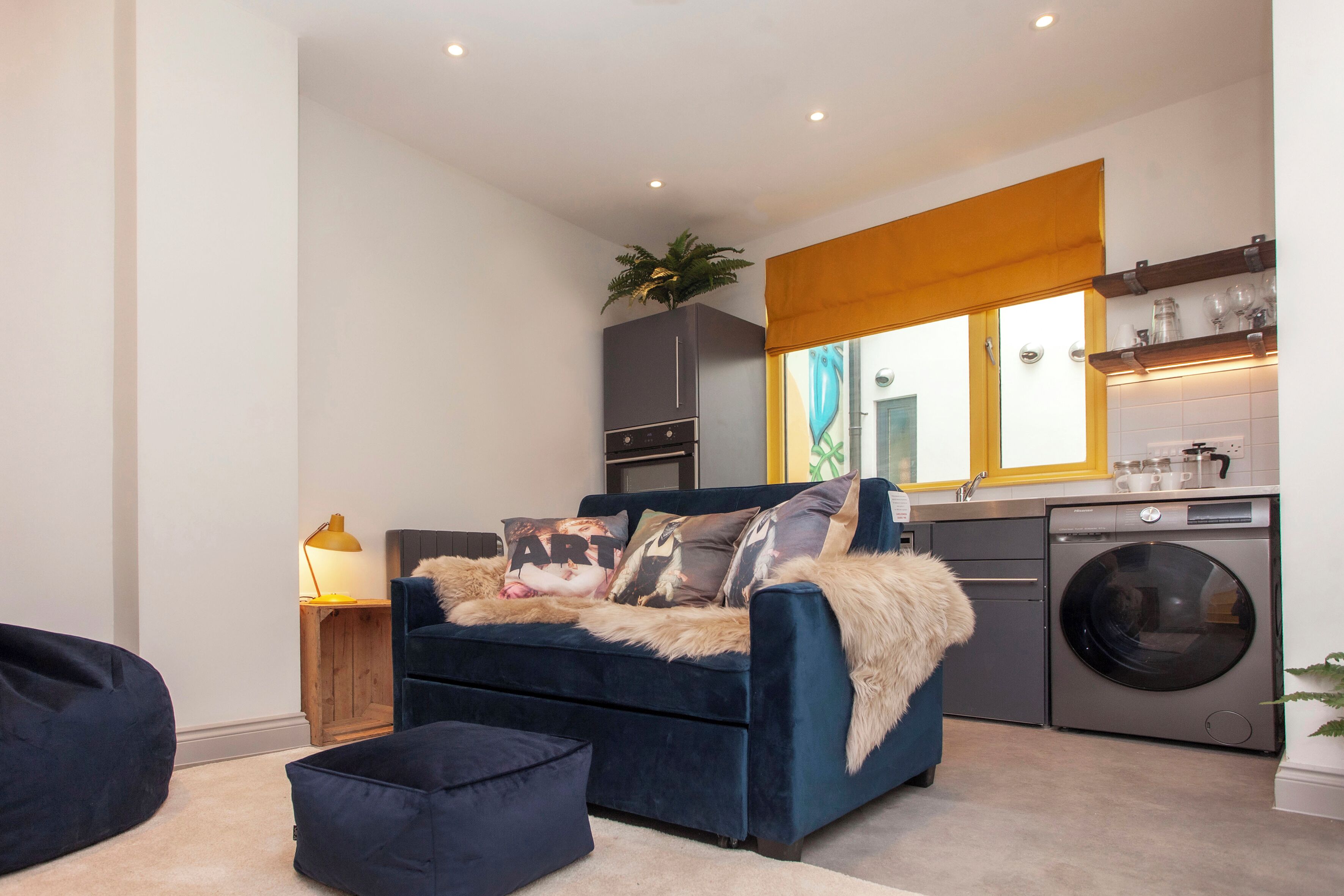 A compact living space with a dark blue sofa, a small wooden side table with a lamp, a washing machine, and a window with a yellow blind.