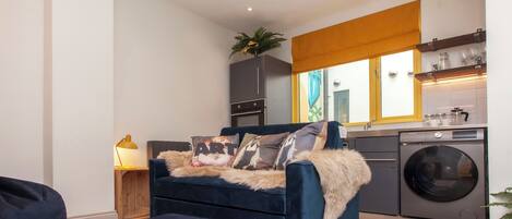 A compact living space with a dark blue sofa, a small wooden side table with a lamp, a washing machine, and a window with a yellow blind.