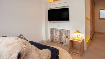 A modern hotel room with a large flat-screen TV mounted on the wall, a wooden sideboard, a small wooden table with a lamp, and a plush fur throw on the sofa.