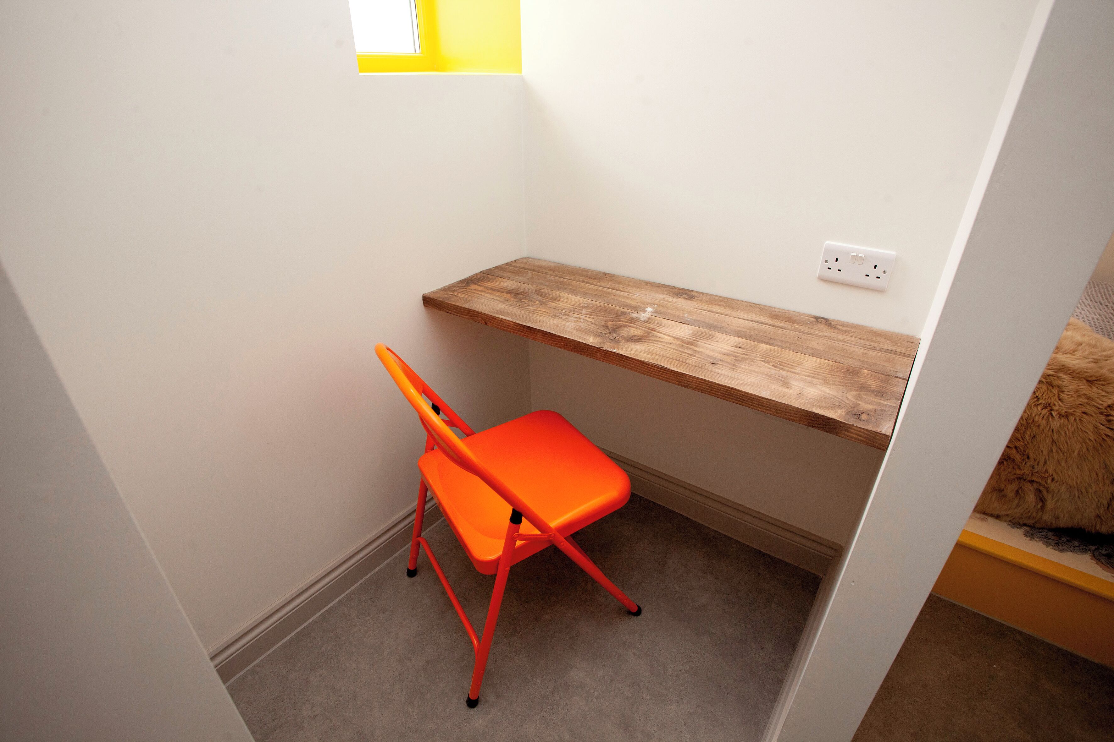A small, narrow room with a wooden shelf, a single orange chair, and a power outlet on the wall.