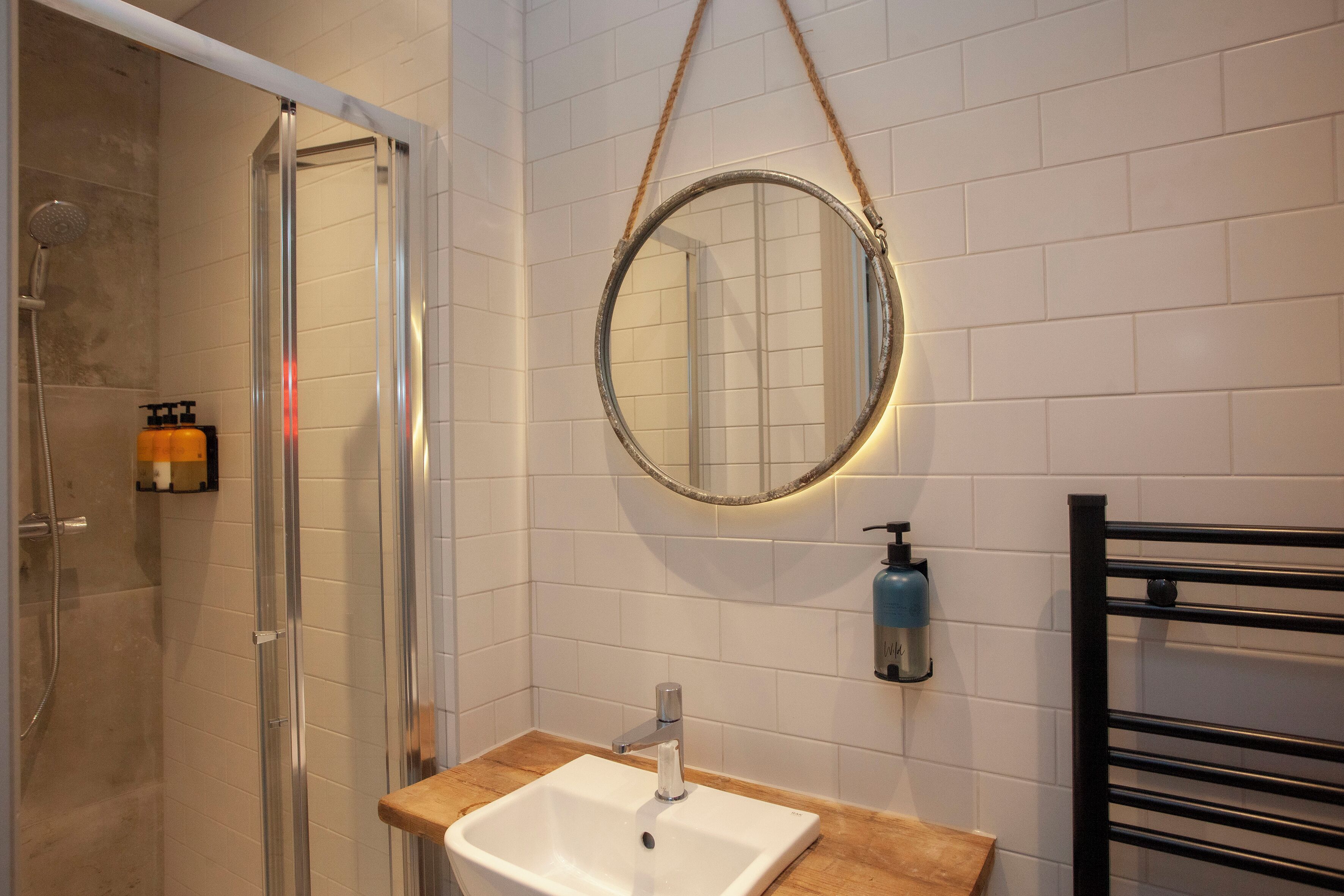A bathroom with a glass shower enclosure, a white sink with a wooden countertop, a round mirror with a rope handle, and a black towel rack.