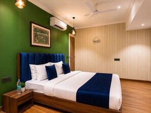 Una habitación de hotel con cama, mesita de noche, botella de agua, un cuadro enmarco, aire acondicionado, ventilador de techo y un letrero de bienvenida.