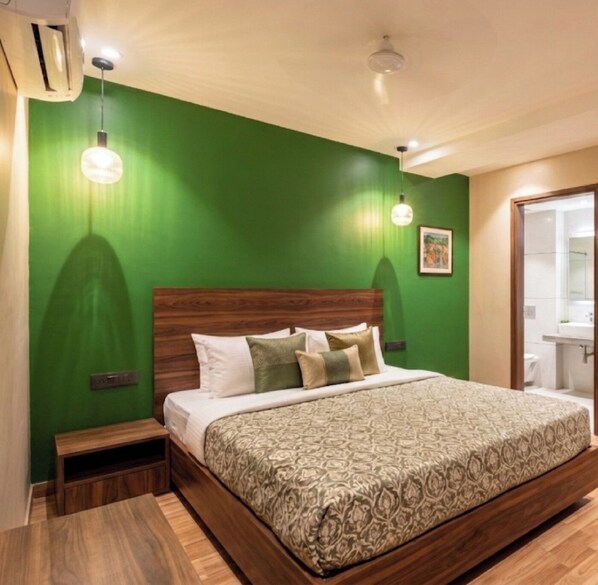 Un dormitorio con pared verde, cabecero de madera, colcha estampada, mesitas de noche y un baño a la vista.