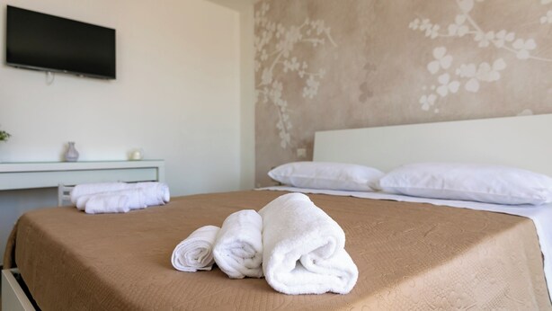 A hotel room with a bed, a flat-screen TV mounted on the wall, a bedside table with a vase, and a floral-patterned wall.