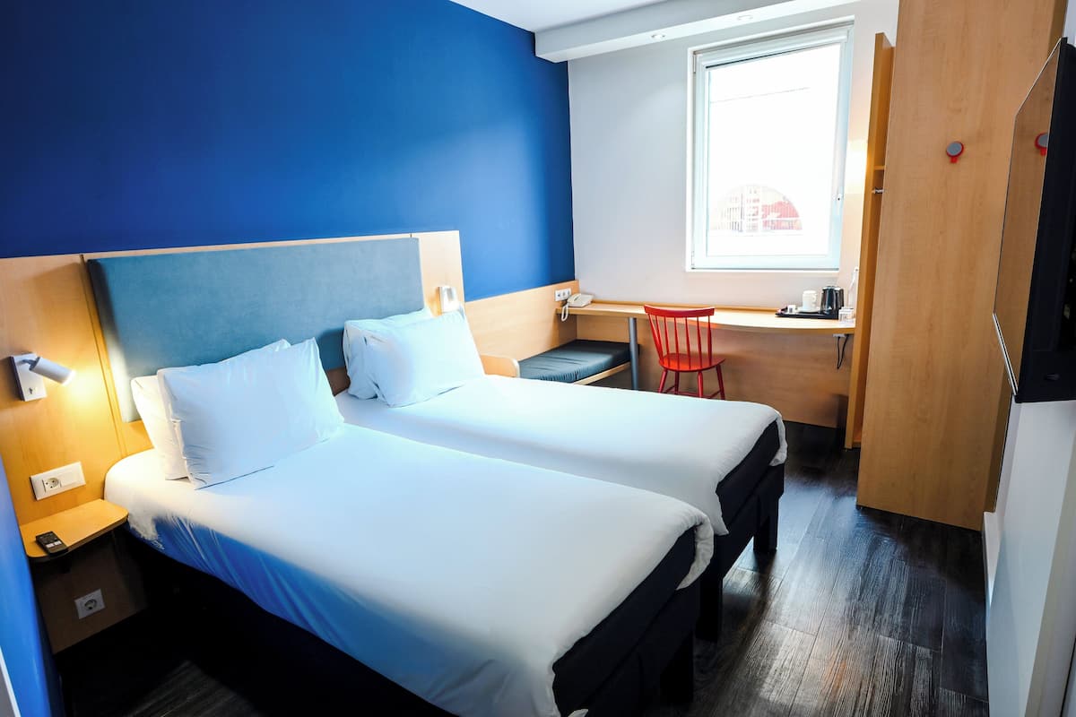 Une chambre d’hôtel avec deux lits, un bureau, une chaise, une fenêtre et un téléviseur.