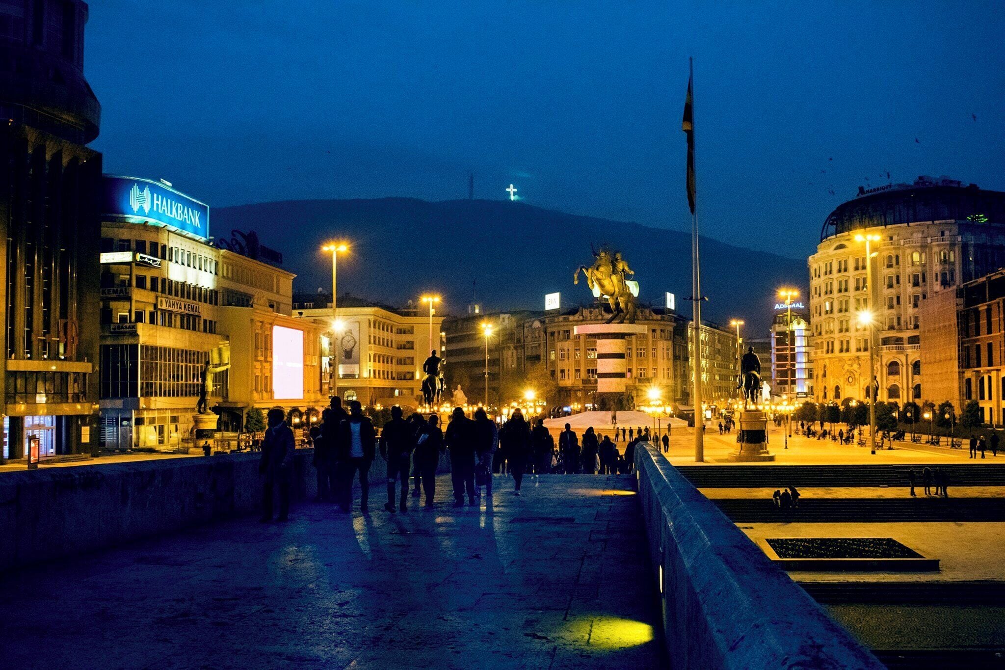 A nighttime city square with a statue, illuminated buildings, and a crowd of people.