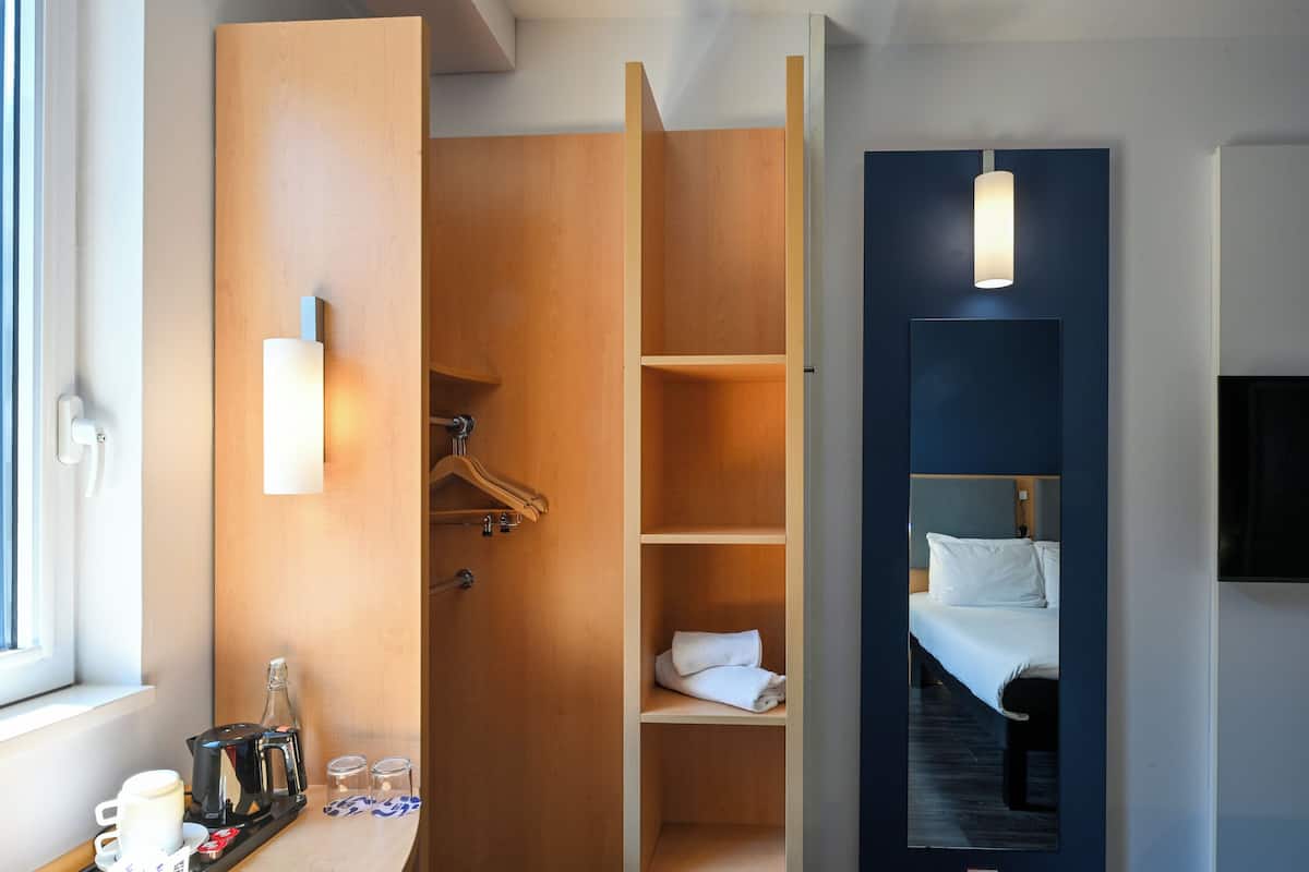 Une chambre d’hôtel avec une armoire en bois, un lit avec une literie blanche, une table de chevet avec une machine à café et un miroir.