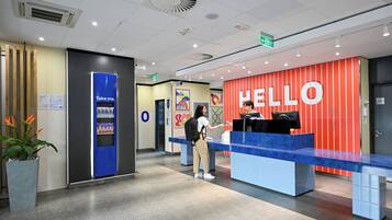 A reception desk with a 'HELLO' sign, a blue counter, a vending machine, and a potted plant.