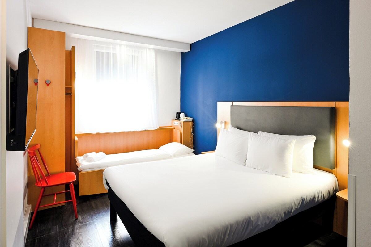 A hotel room with a bed, a chair, a desk, and a window with curtains.