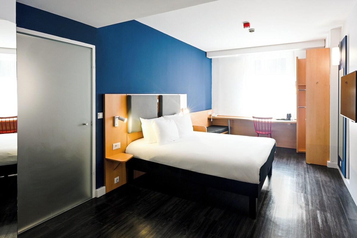 A modern hotel room with a large bed, a desk, and a wardrobe.