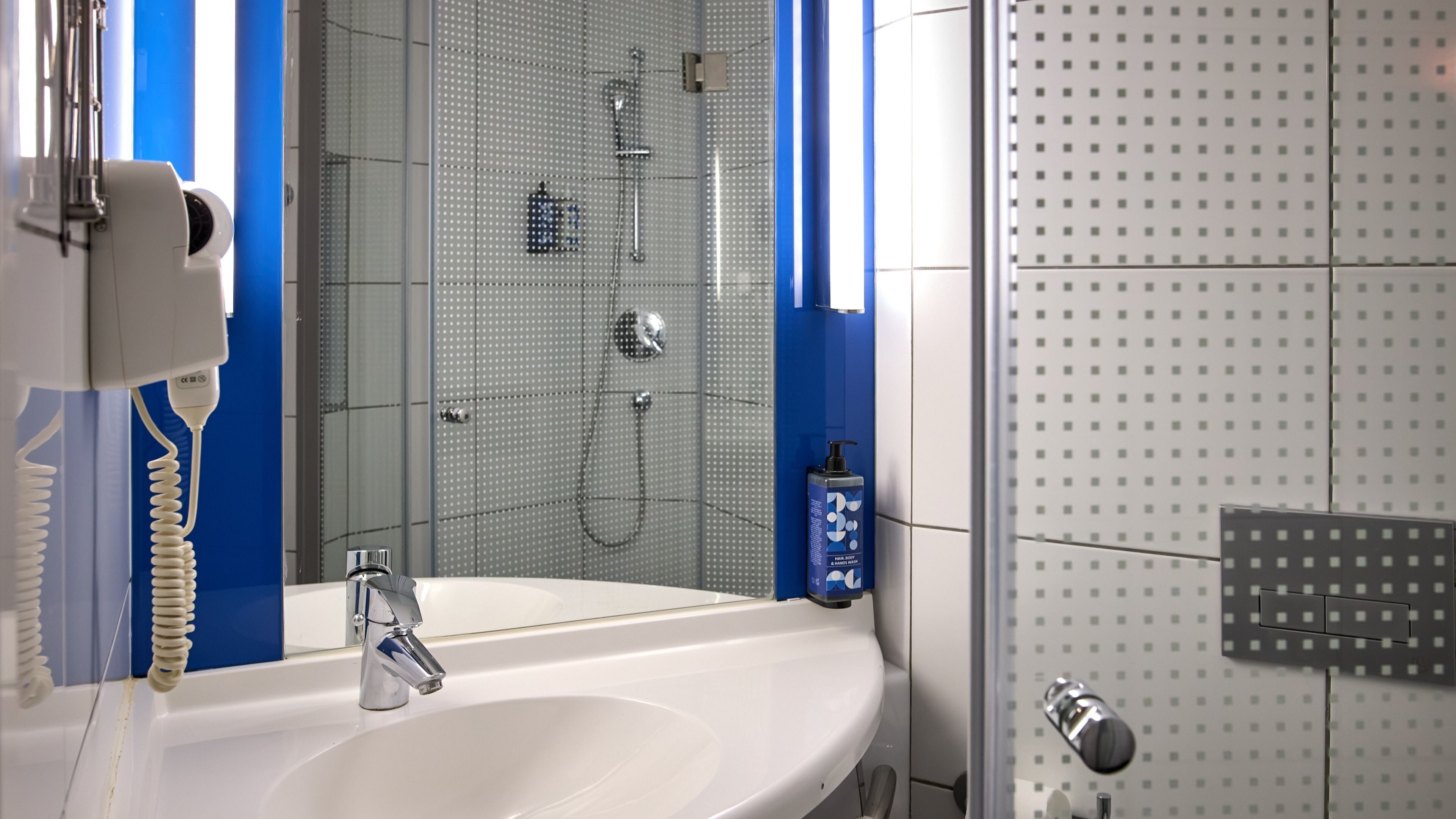 Baño con lavamanos blanco, ducha de mano y zona de ducha con mampara de vidrio.