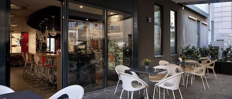 An outdoor café with white chairs and tables, a glass wall with artwork, and a view of the street.