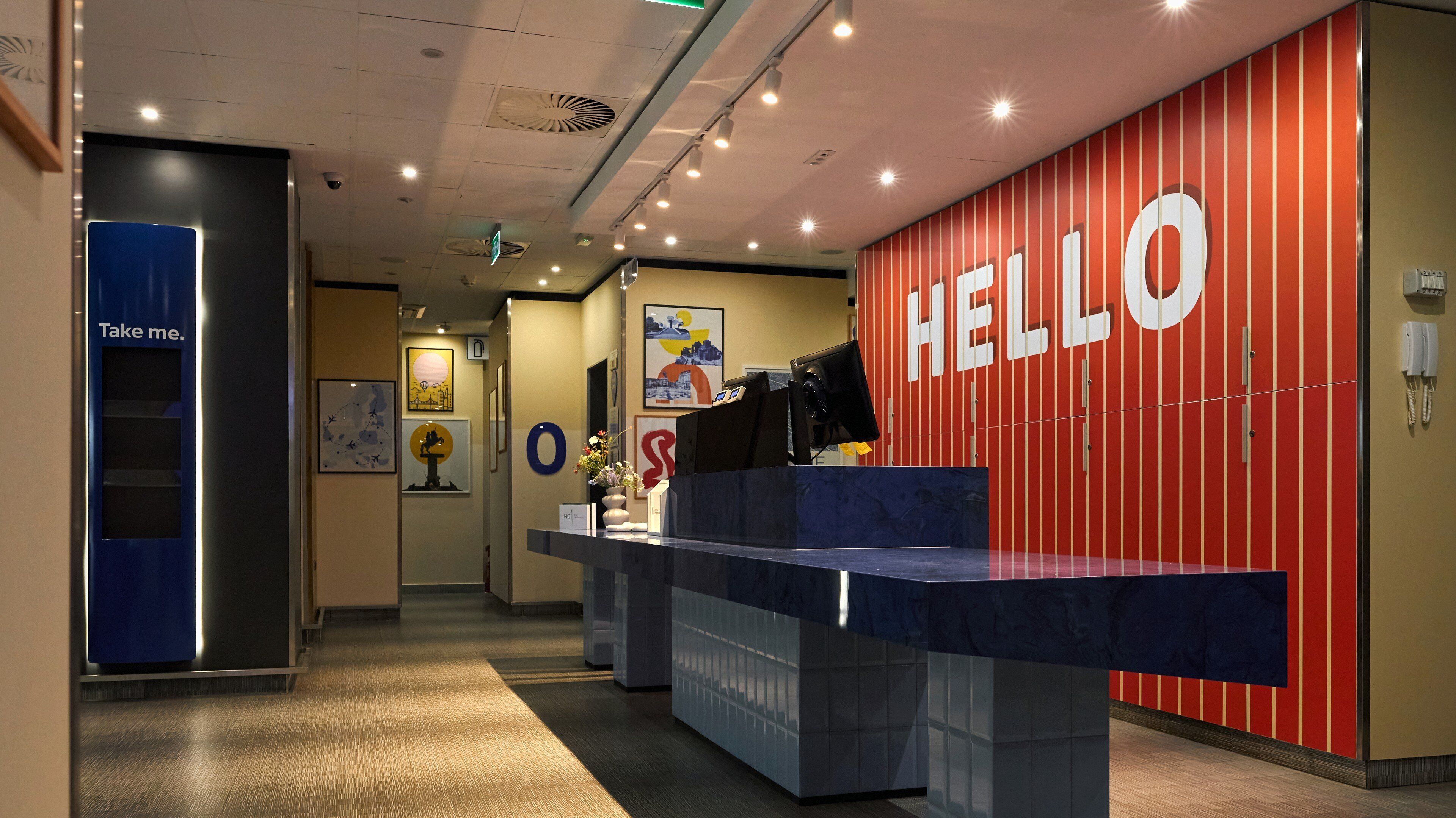 A reception desk with a 'HELLO' sign, a 'Take me' sign, and framed artwork on the walls.
