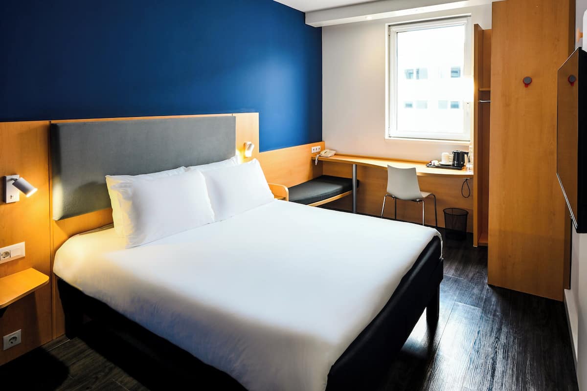 Une chambre d’hôtel avec un lit, une table de chevet, un bureau, une chaise et une fenêtre.