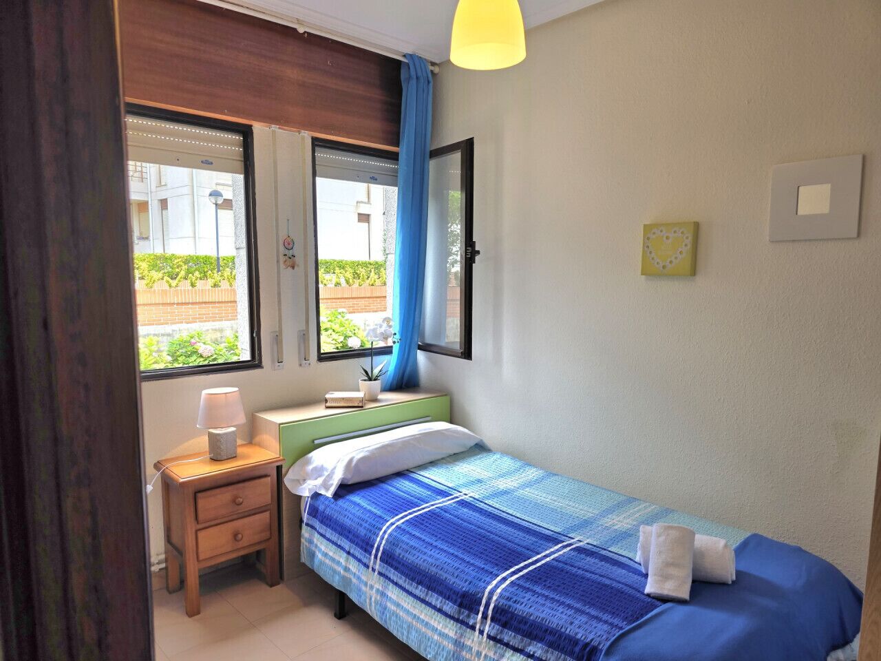 A bedroom with a bed, a nightstand, a lamp, a window with curtains, and a view of the outside.