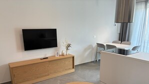 Ein modernes, minimalistisches Hotelzimmer mit einem Wand-Fernseher, einem Holztischchen mit Fernbedienung, einem kleinen Topfpflanzen und einem Essbereich mit weißem Tisch und Stühlen.