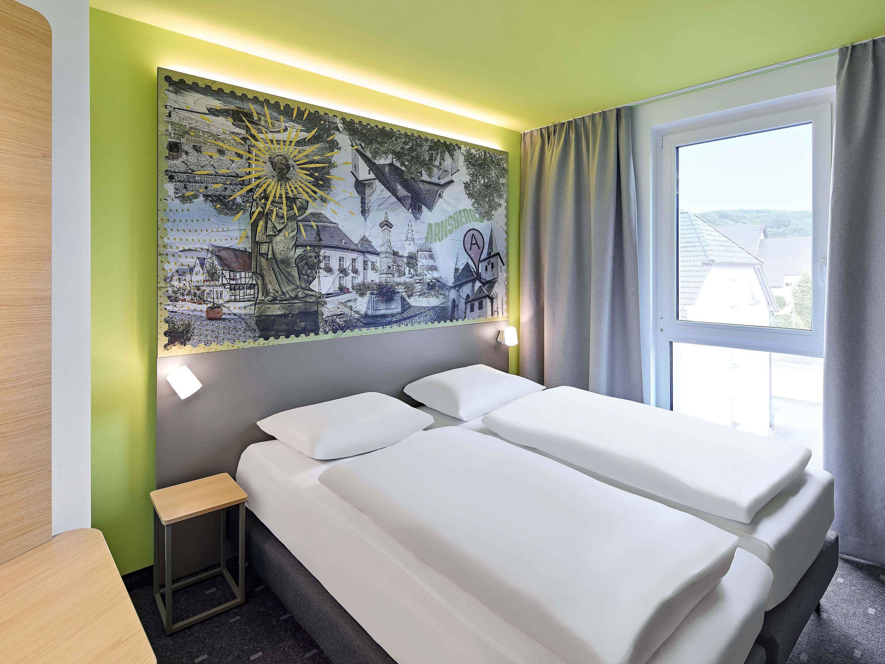 Een hotelkamer met een bed, een klein tafeltje, een wandlamp en een raam met gordijnen.