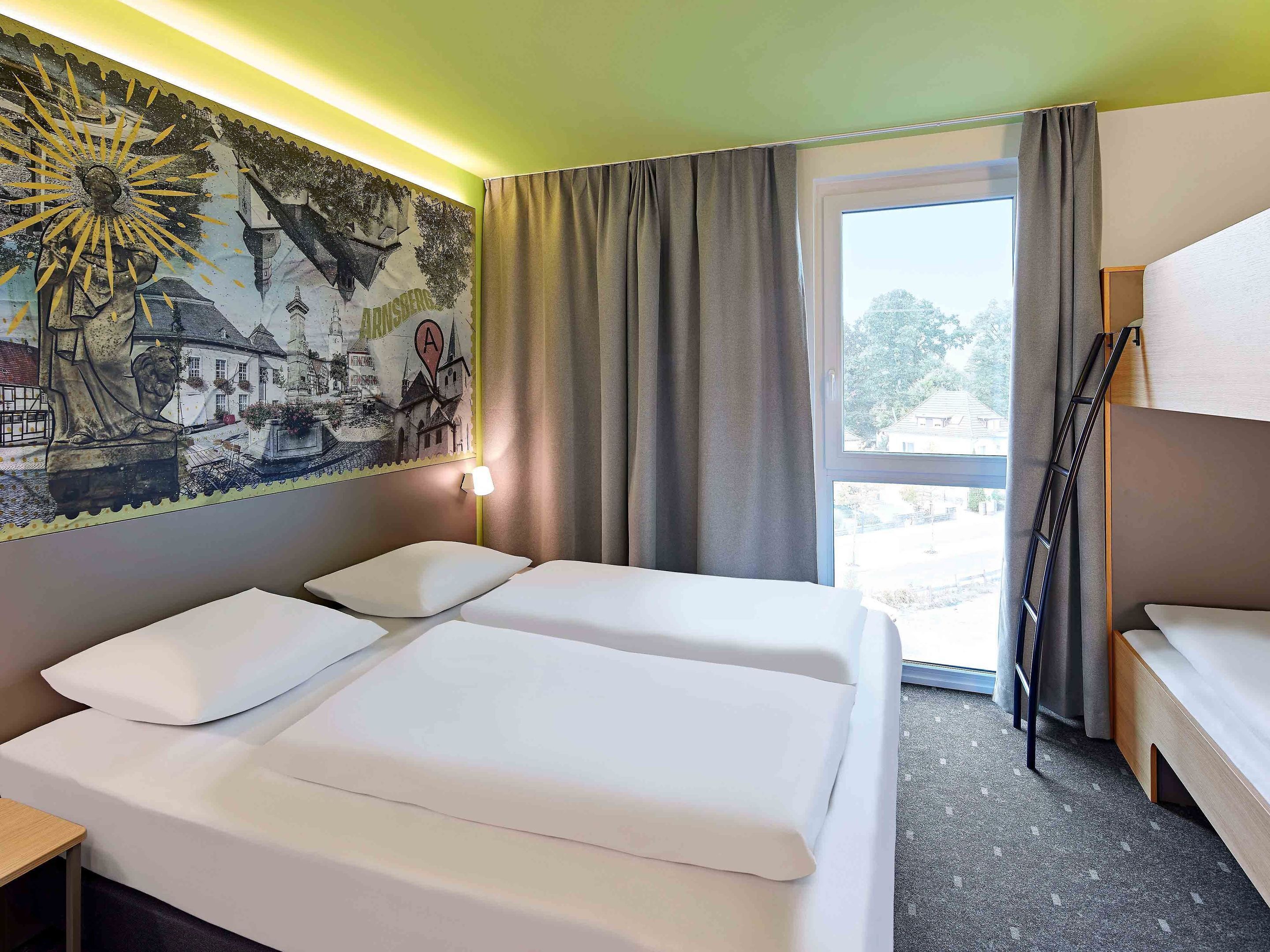 A hotel room with two beds, a mural on the wall, a window with curtains, and a view of the outside.
