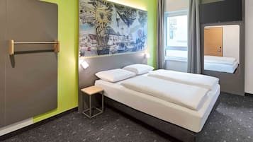 Quarto de hotel com cama, uma mesinha, um quadro na parede e janela com cortinas.