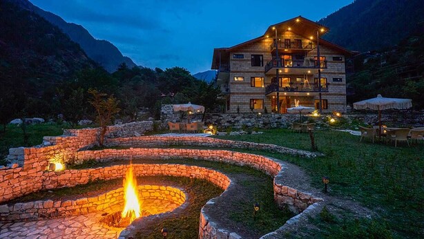 Un espacio al aire libre con paredes de piedra y una chimenea central, rodeado por un sendero de piedra, con un edificio de varios pisos y luces exteriores al fondo.