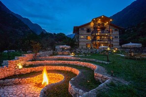 A stone-walled outdoor area with a central fire pit, surrounded by a stone pathway, and a multi-story building with outdoor lighting in the background. - Mashru Riverside Estate (Banjar)