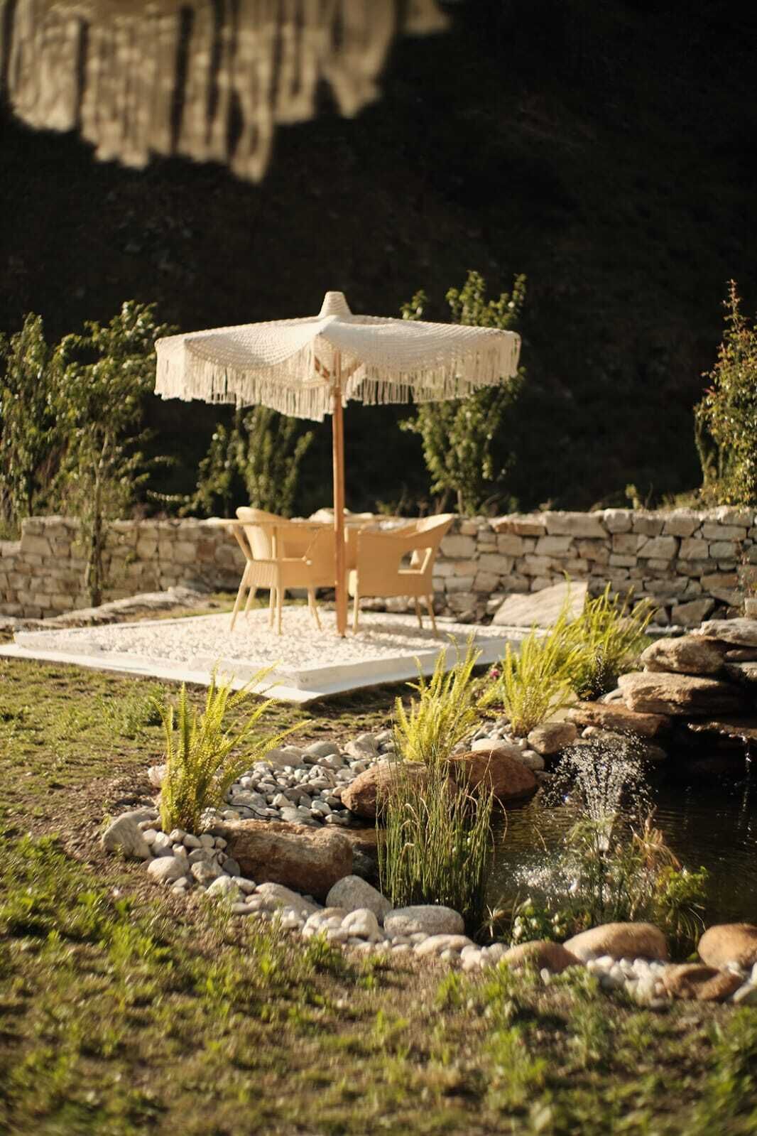 A patio with a table and chairs under a straw umbrella, surrounded by a small pond and rocks.
