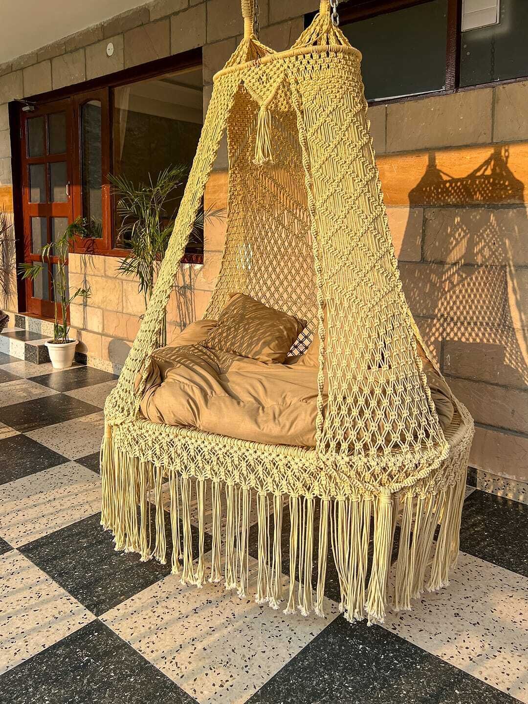 A macrame hanging chair with a canopy, placed on a checkered floor with potted plants nearby.