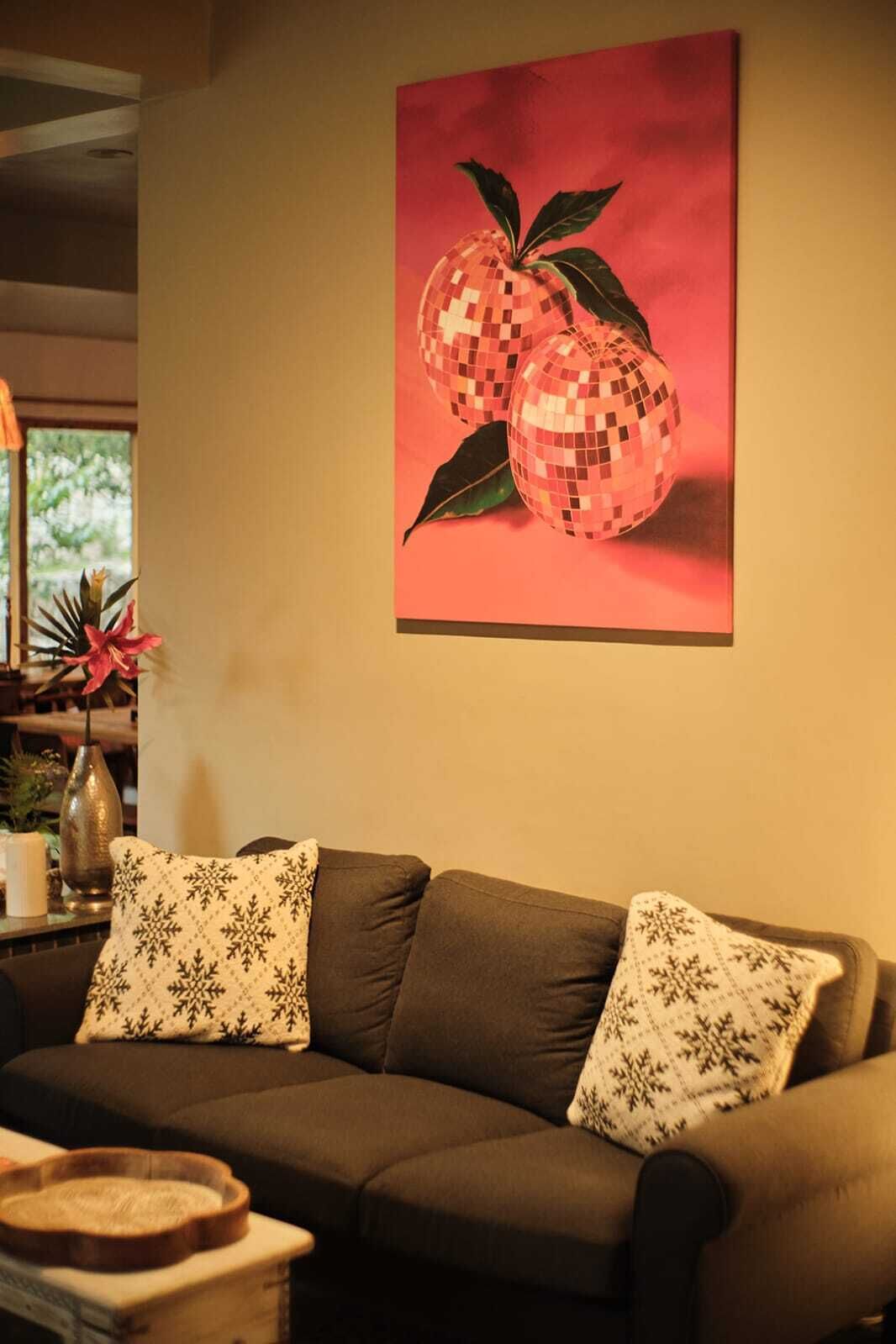 A living room with a brown sofa, two patterned pillows, and a wall-mounted painting of a fruit.