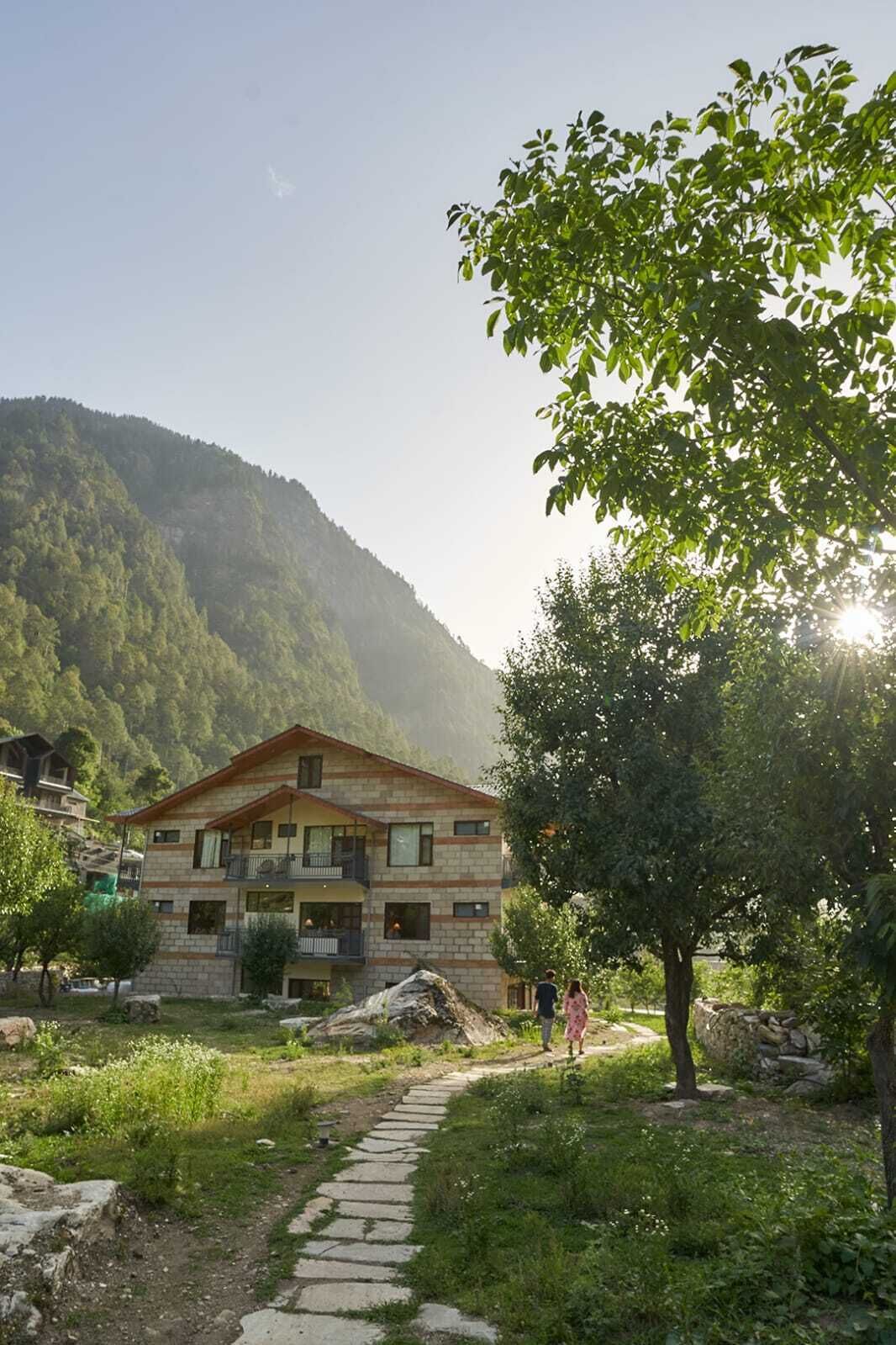 A two-story building with a stone facade and a balcony, surrounded by greenery and a mountain backdrop.