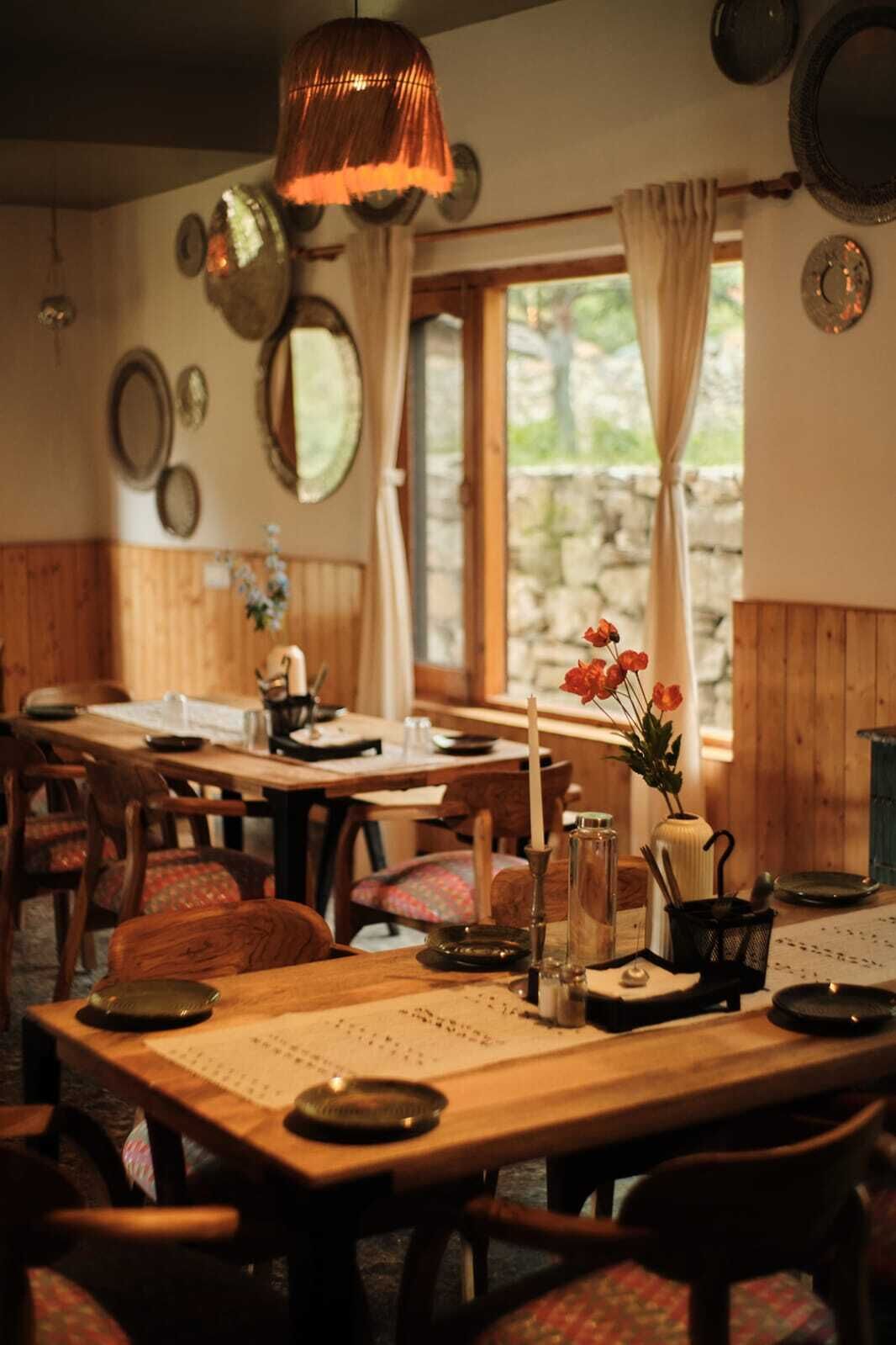 A cozy dining area with wooden tables and chairs, a hanging copper light fixture, and a window with curtains.