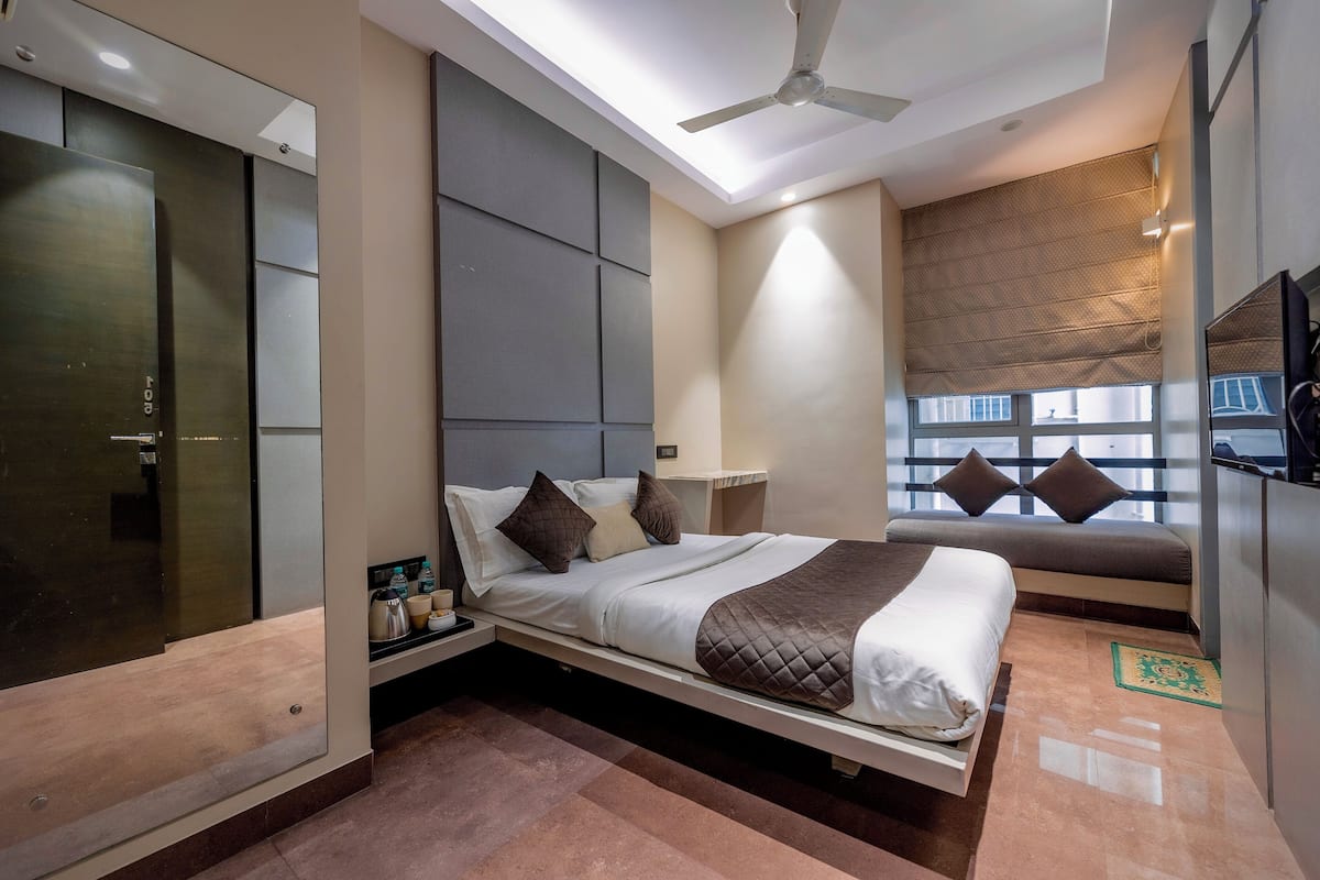 A modern hotel room with a bed, a ceiling fan, a window with blinds, and a TV on the wall.