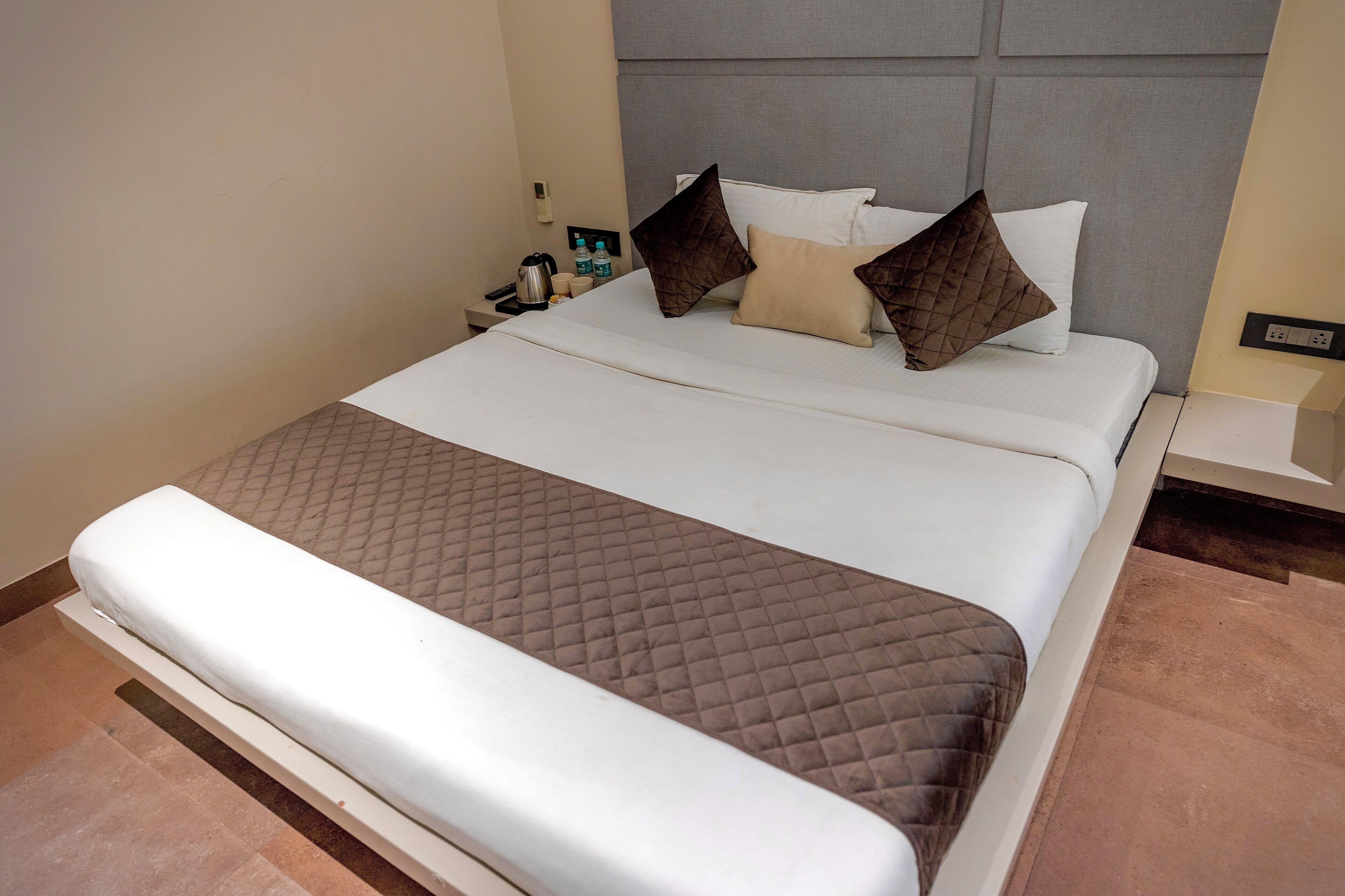 A neatly made bed with a quilted blanket, a headboard with a padded design, and a bedside table with a water bottle and a cup.