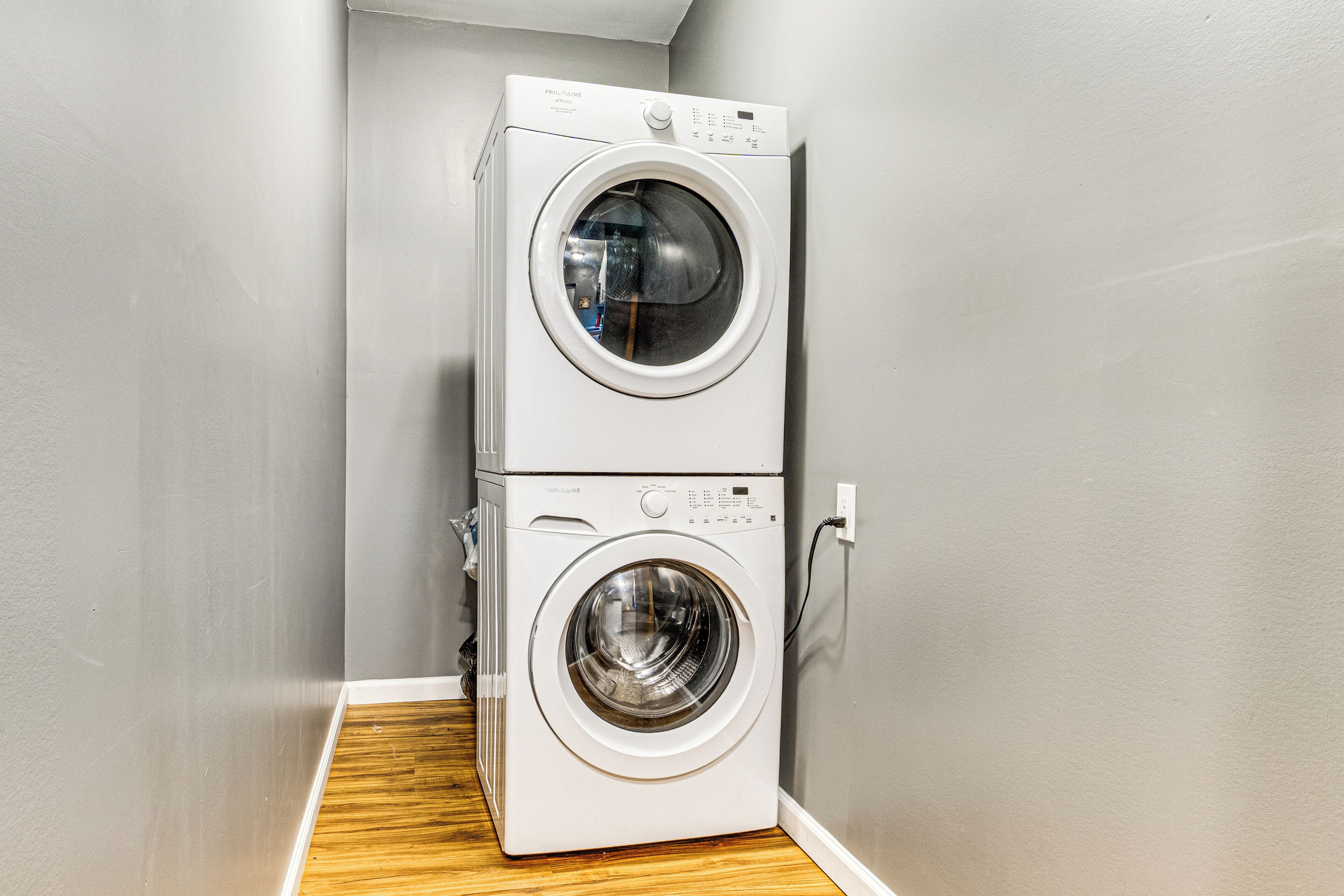 A stacked washer and dryer unit in a corner with a wooden floor and grey walls.