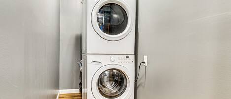 A stacked washer and dryer unit in a corner with a wooden floor and grey walls.