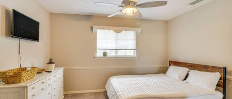 Un dormitorio con cama, ventilador de techo, televisión, cómoda y ventana con persianas.