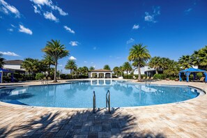 A clear blue swimming pool with a tiled deck, surrounded by palm trees and a building with a covered patio. - Pool, Game Room & More! Resort Home 9 Mi to Disney (Kissimmee)