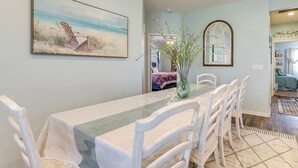 A dining area with a long table, white chairs, a beach-themed painting, and a vase with branches.
