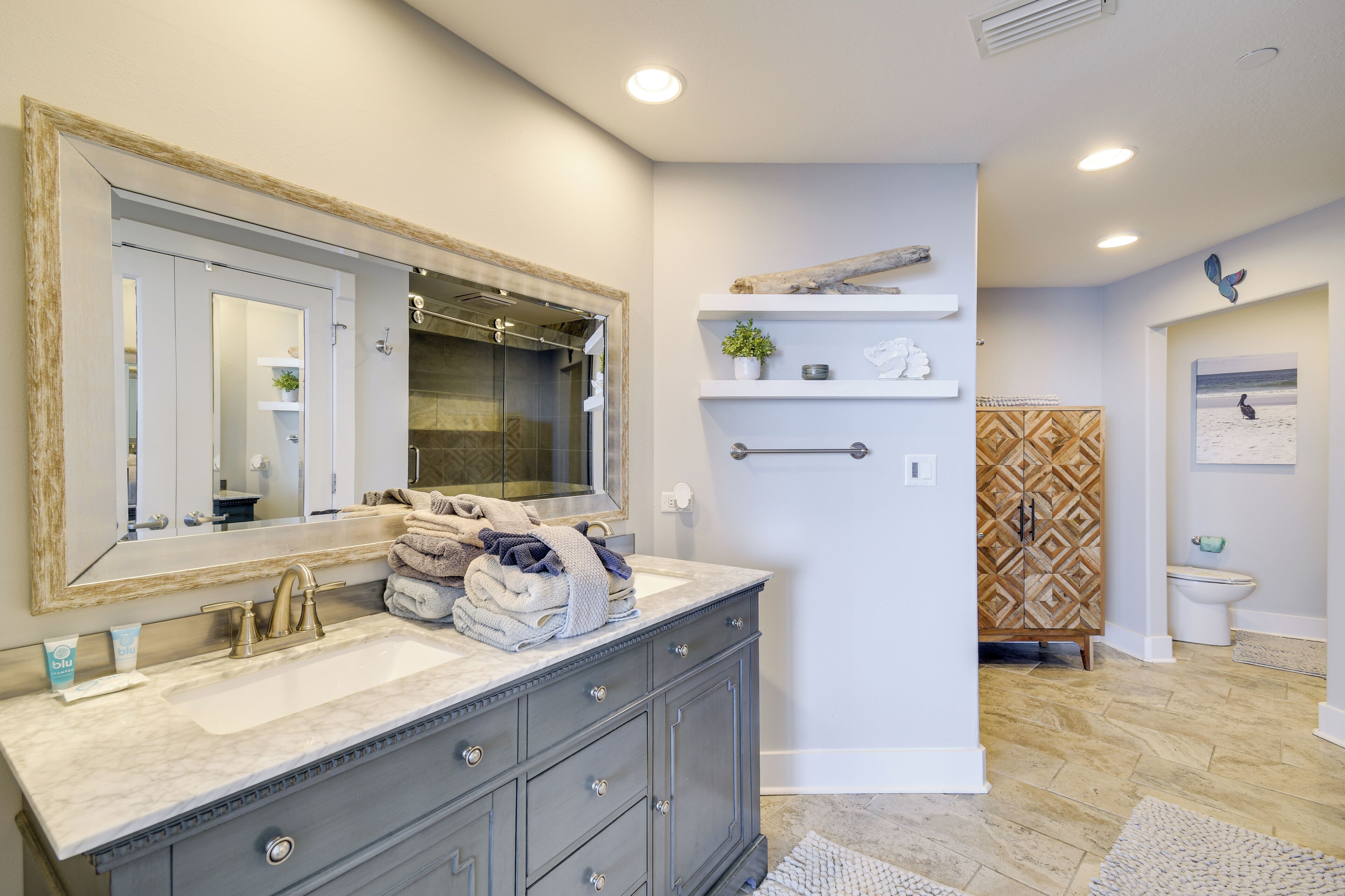 A bathroom with a large mirror, dual sinks, and a vanity with drawers. There are towels stacked on the vanity and a shelf with decorative items.