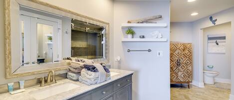 A bathroom with a large mirror, dual sinks, and a vanity with drawers. There are towels stacked on the vanity and a shelf with decorative items.