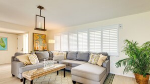 A living room with a sectional sofa, a coffee table, a hanging light fixture, and a large window with blinds. - Riverfront Home w/ Private Dock on Merritt Island! (Merritt Island)