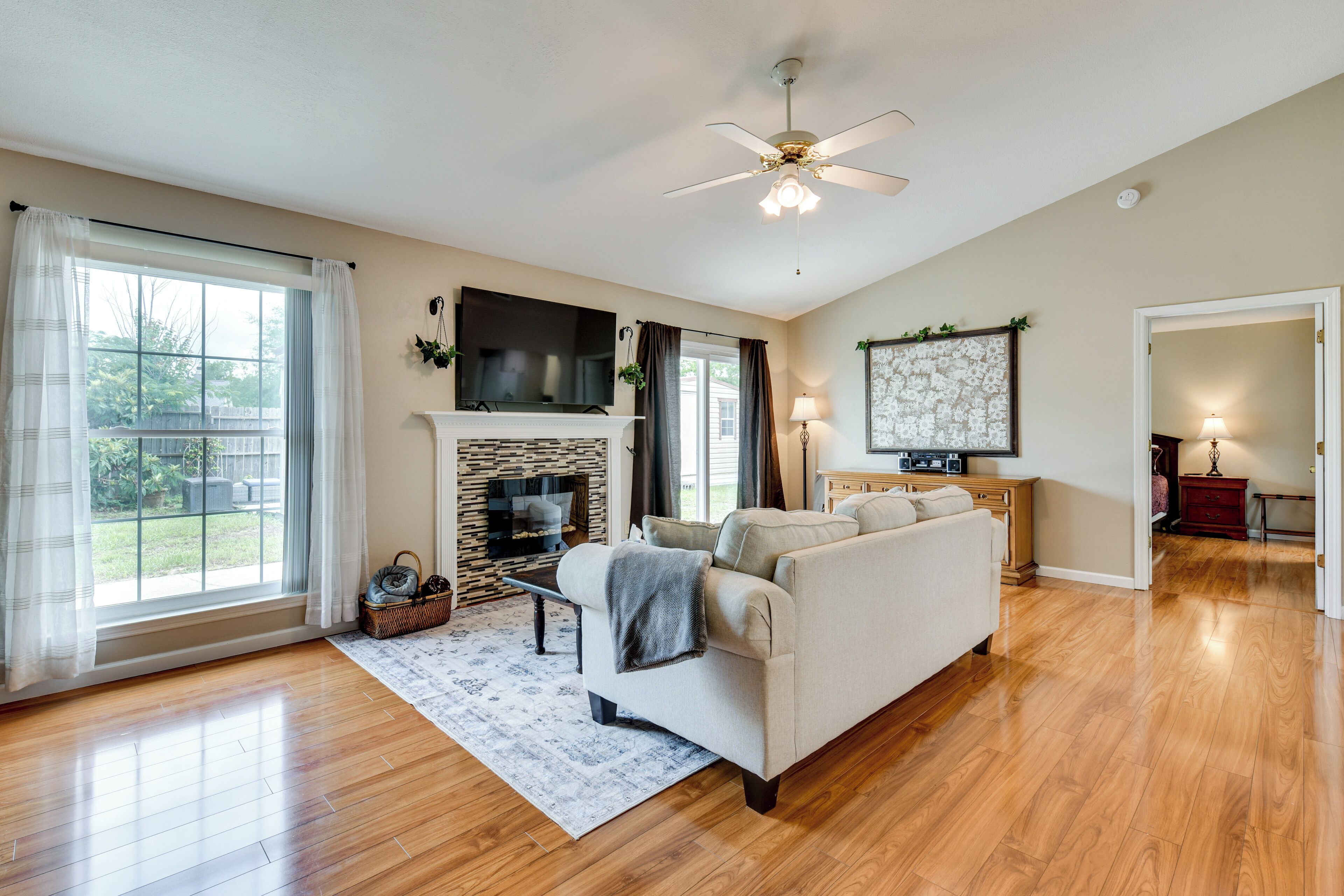 A spacious living room with a fireplace, a sofa, a ceiling fan, and a large window with curtains.
